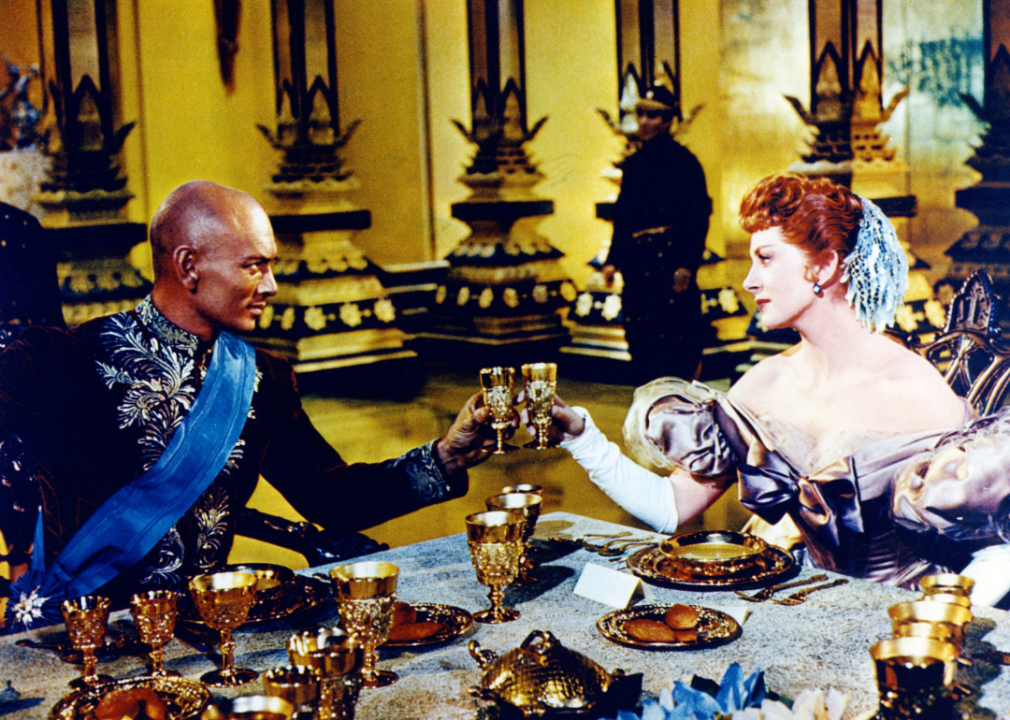 Yul Brynner and Deborah Kerr in costume, seated at a dining table.