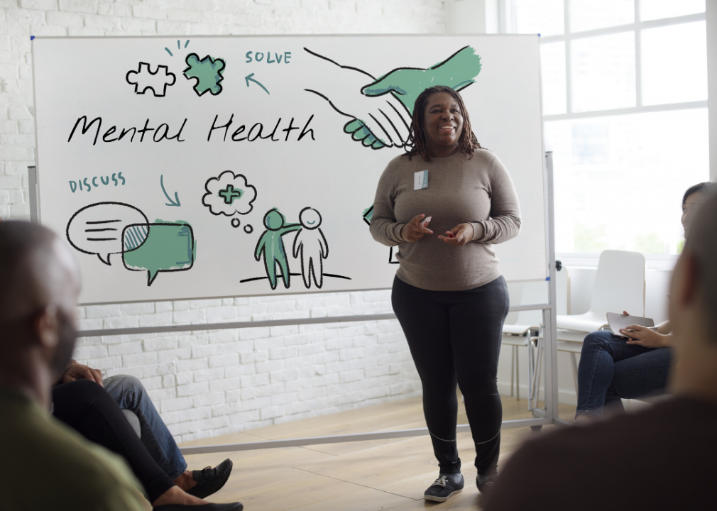 A smiling woman stands in front of a white board with the words mental health, and solve and discuss. 