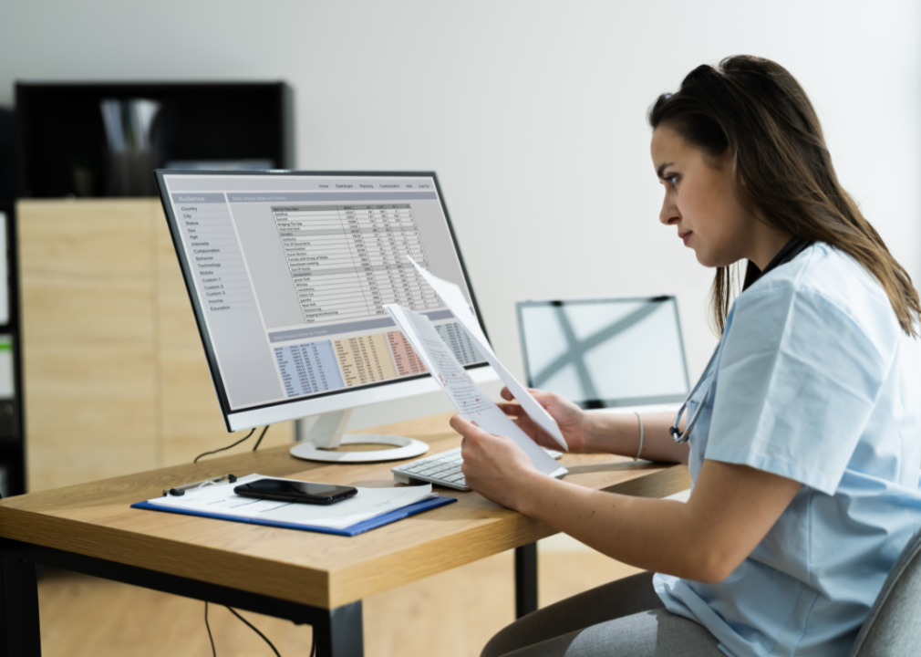 A woman in blue medical scrubs sits at a computer and looks at paperwork.