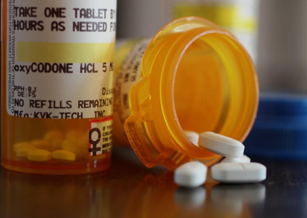 A close up of two orange bottles on a table, one standing upright with its prescription label for oxycodone visible, and the other on its side with the top off and a few white pills spilling out.