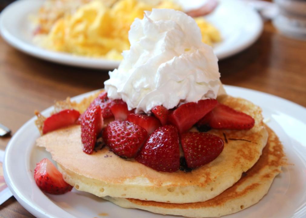 Pancakes with whipped cream and strawberries with a plate of scrambled eggs in the background.