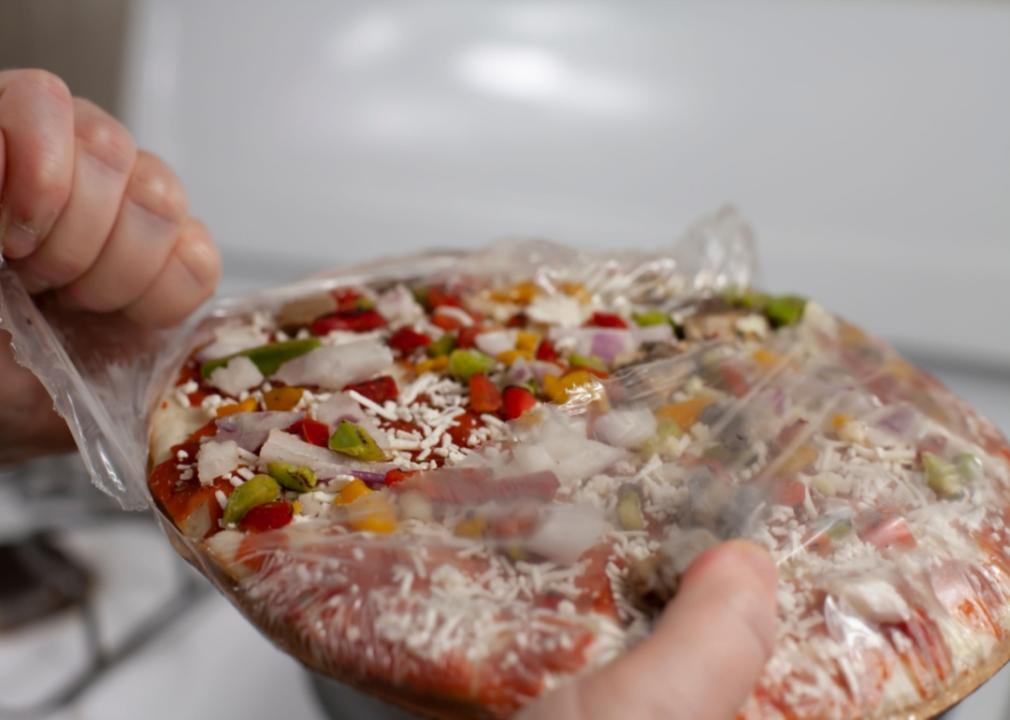 A close up of hands tearing the plastic off a frozen pizza.