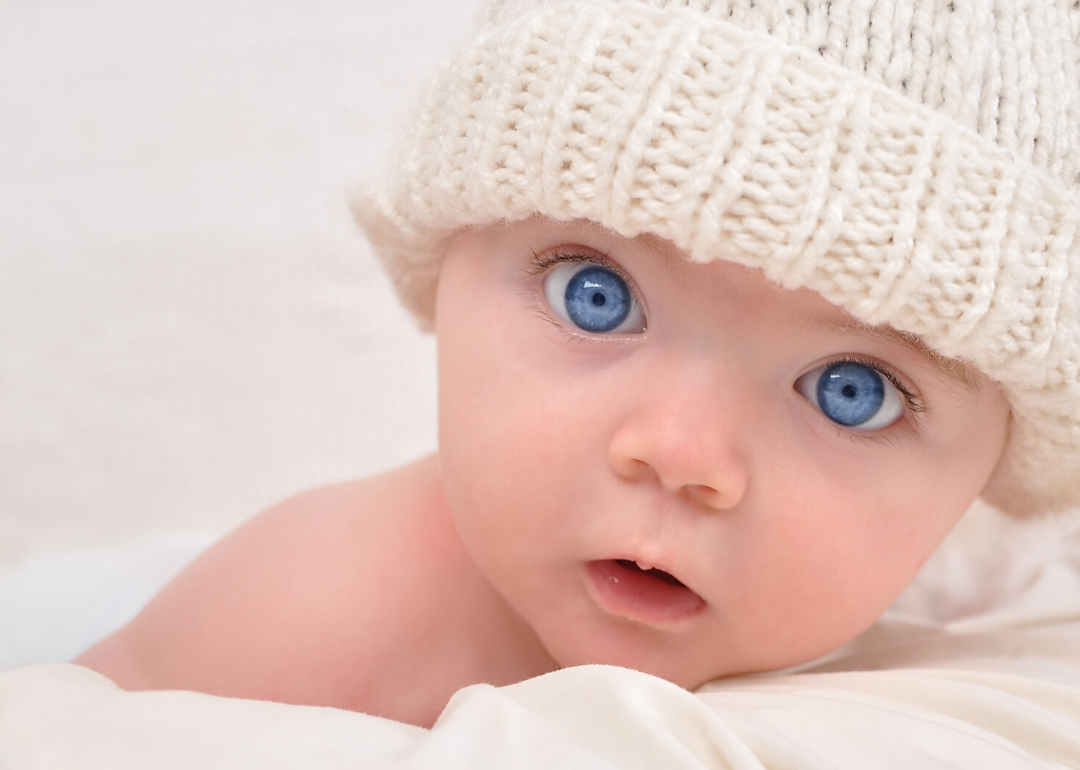 Close up of a baby with blue eyes and white knitted cap on.