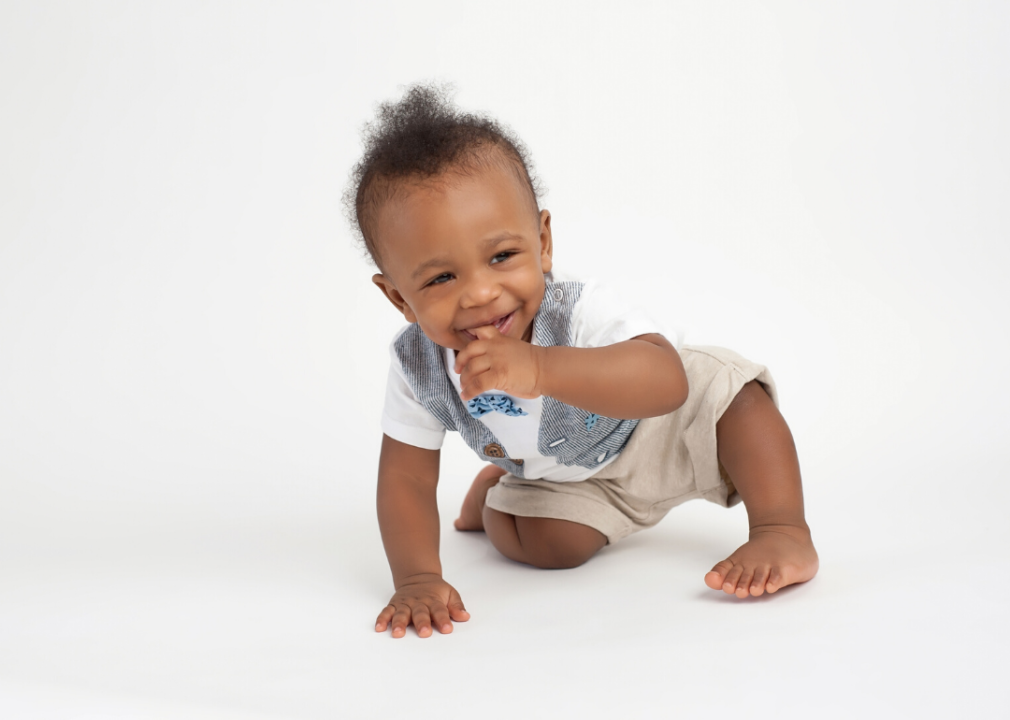 A smiling baby boy sitting on a white, seamless background with his finger in his mouth.