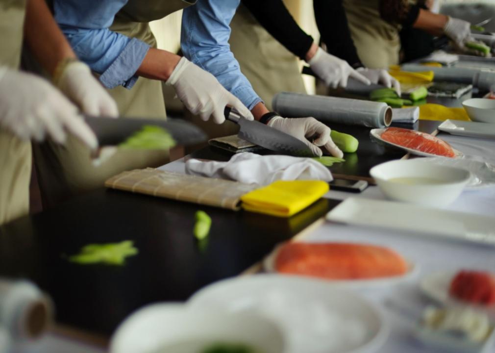 A close up of several peoples' gloved hands slicing ingredients for making sushi.