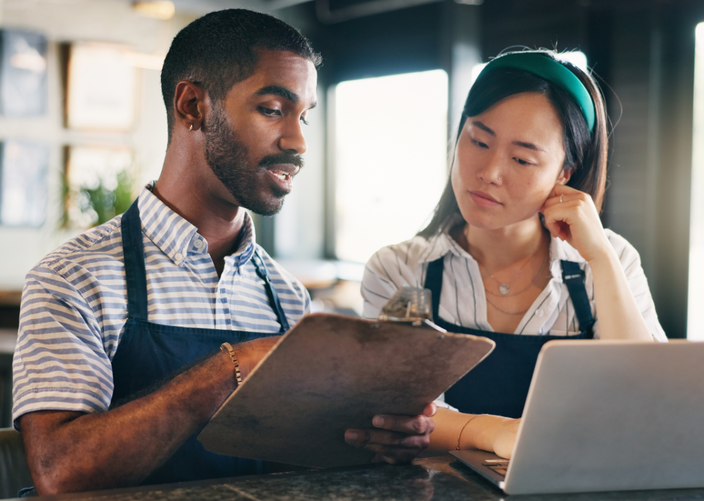 A manager training an employee in a shop with them both looking at a clipboard in front of a laptop.