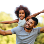 Father carrying daughter on shoulders in park.