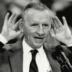 Ross Perot speaks with hands to his ears.