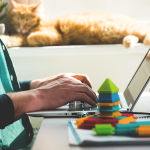 Person working remotely with children’s blocks on desk and a cat in background.