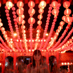 Red lanterns line the ceiling above a woman with her eyes closed and her hands in prayer. 