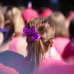 Woman, photographed from behind, stands in a crowd wearing a pink pony tail with a pink ribbon as a symbol for fighting cancer. She is surrounded by ladies wearing pink, all raising money to fight cancer
