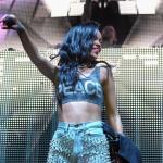 Rihanna wears a crop top that reads "Peace" and holds the mic up to the audience while performing on stage.