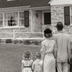A family of five in the 1950s with their backs to the camera standing on the lawn looking at a fieldstone house.