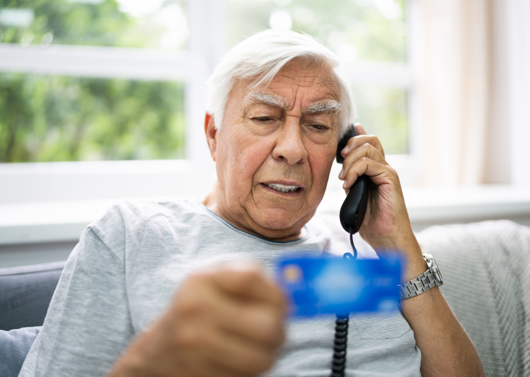 Senior man sitting on a couch reading off his credit card information on the phone.