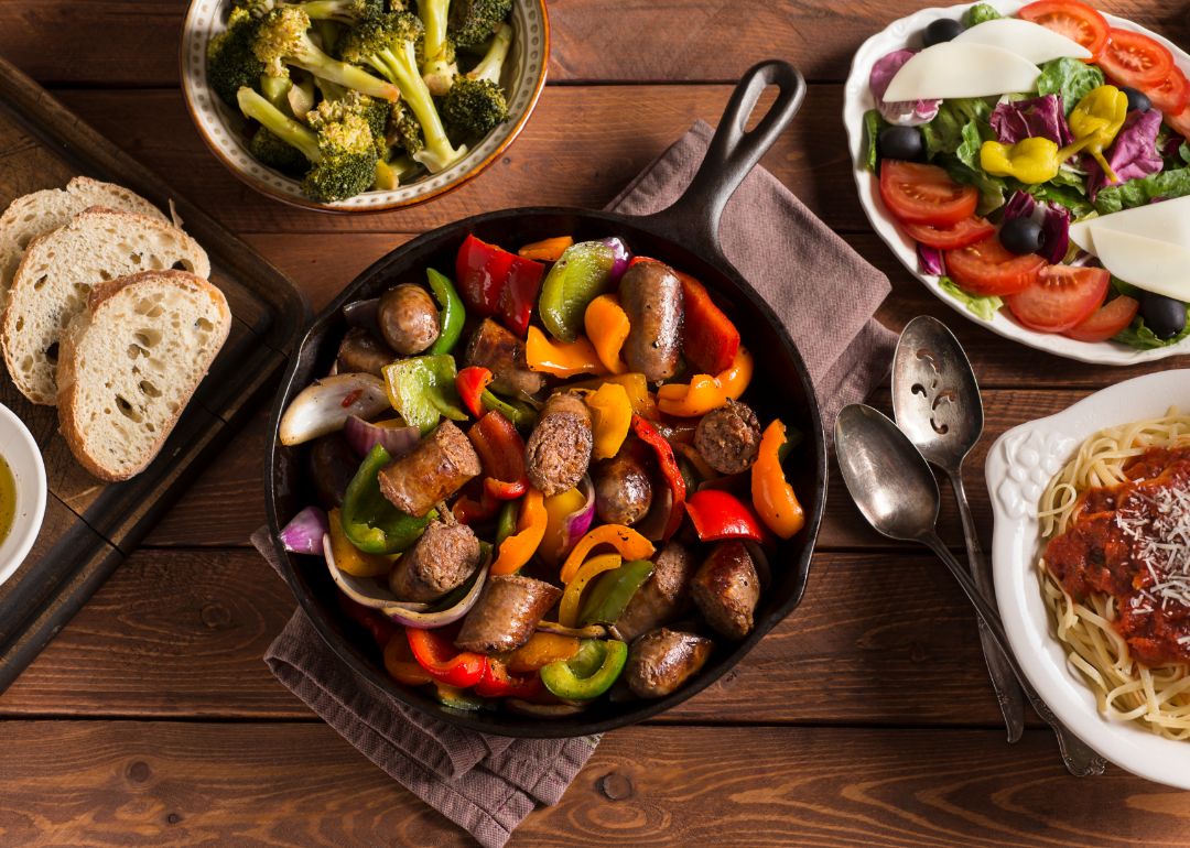 Sausage, peppers, and onions featured at the center of a spread of Italian food on a wooden table.
