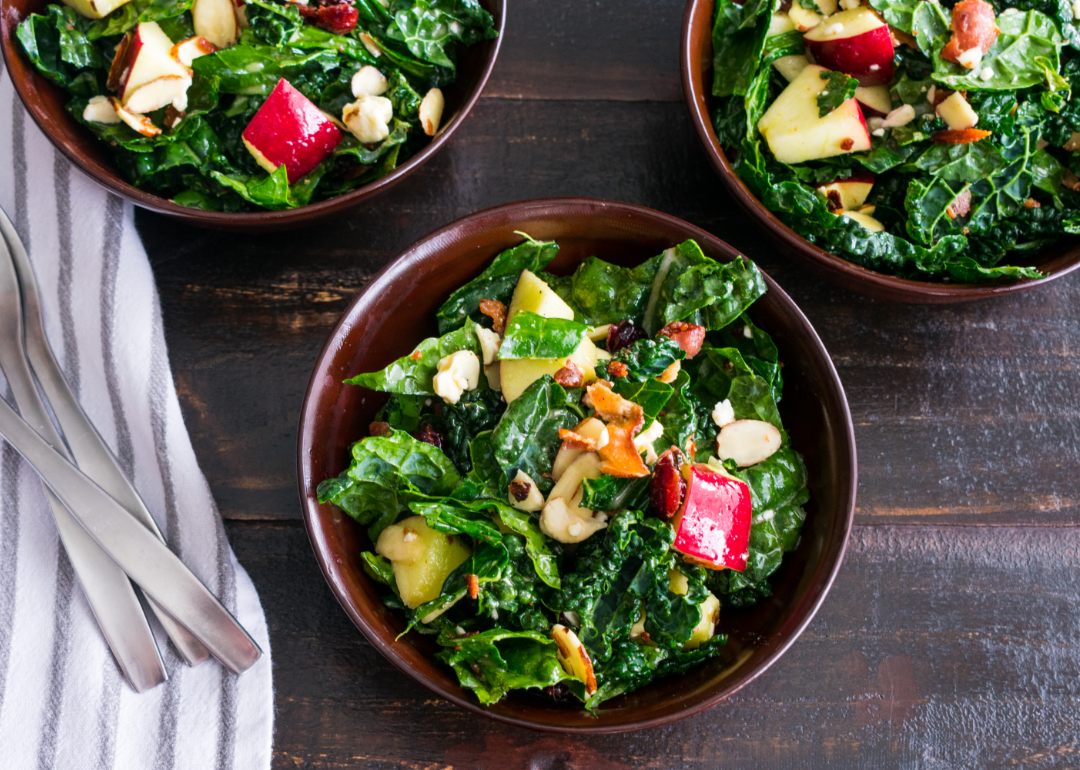 Apple cranberry bacon kale salad served in three bowls on a wooden table.