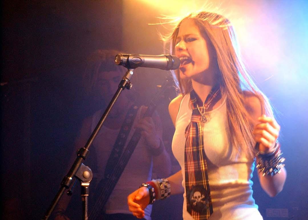 Avril Lavigne sings while wearing a white tank top and a plaid tie.
