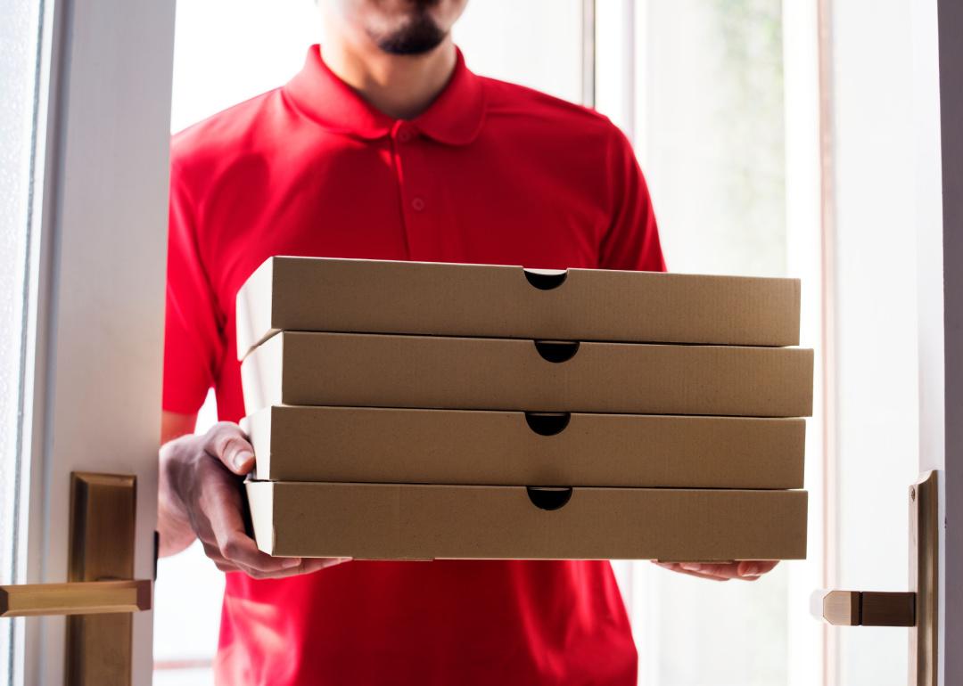 Delivery person in red shirt standing at a door with four pizza boxes.