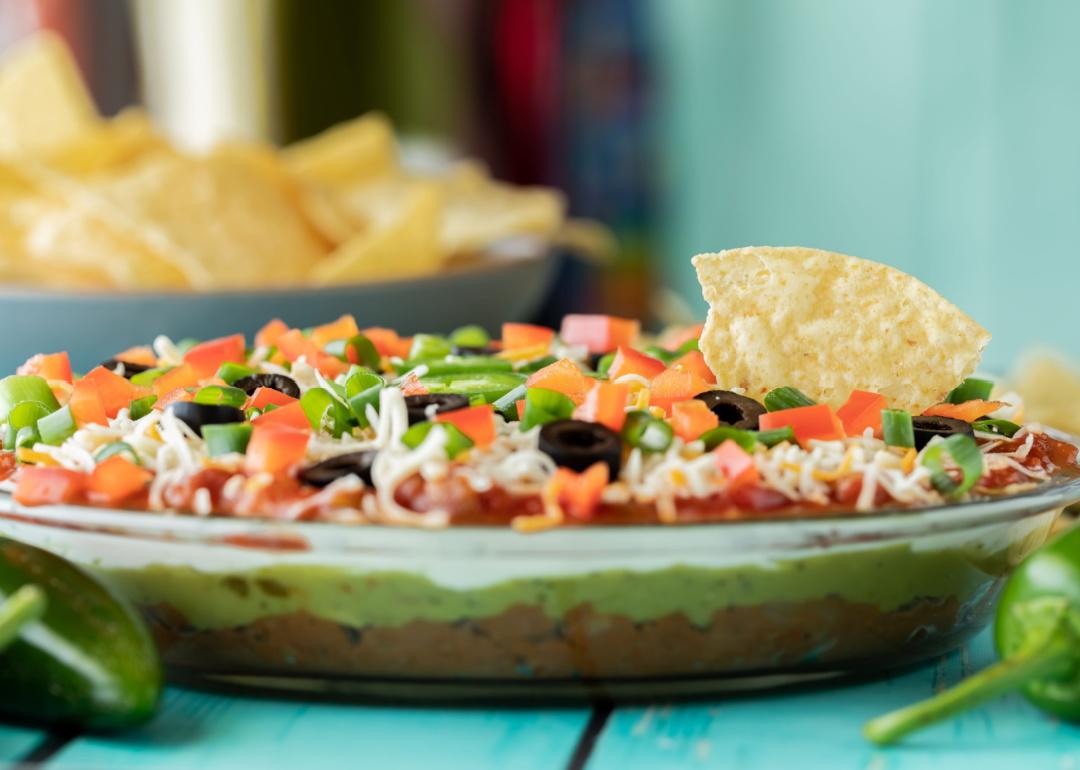 A seven-layer taco dip with one tortilla chip dipped in and a bowl of chips in behind it.