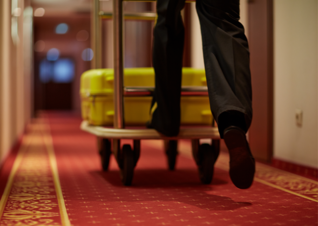 Closeup on legs of bellhop pushing luggage cart with yellow suitcase down a hotel hallway.