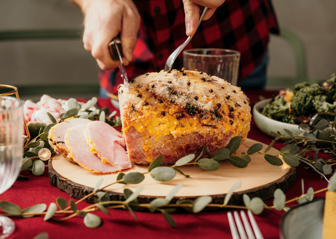 Closeup of person in a flannel shirt cutting into a ham on a Christmas table.