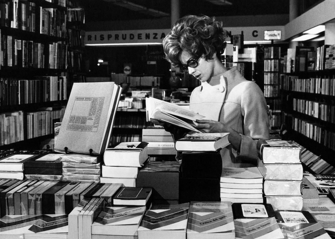 A person with a bouffant and cat-eye glasses leafing through a book at the bookshop in the 1960s.