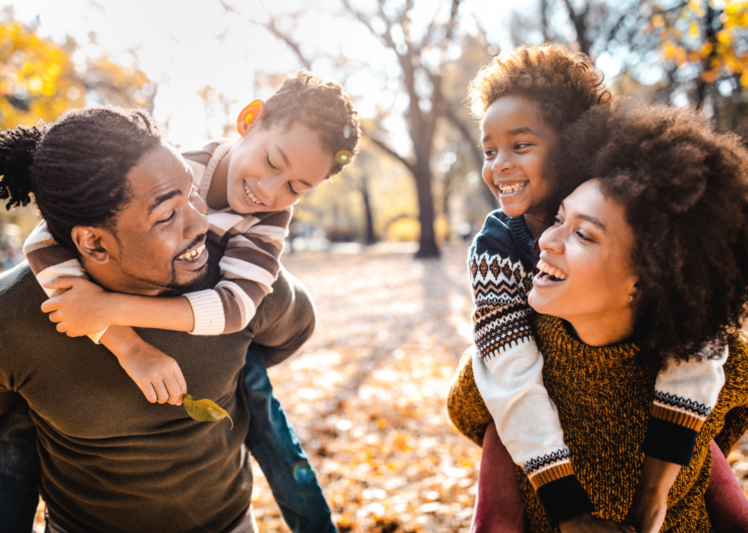 A Black family of four laughing and piggybacking in a park on an autumn day.