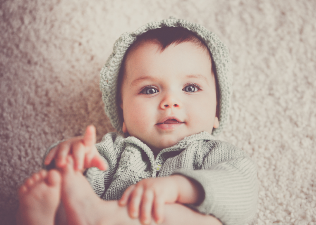Cute baby with brown hair and blue eyes in a matching knit hat and sweater lying on the carpet.