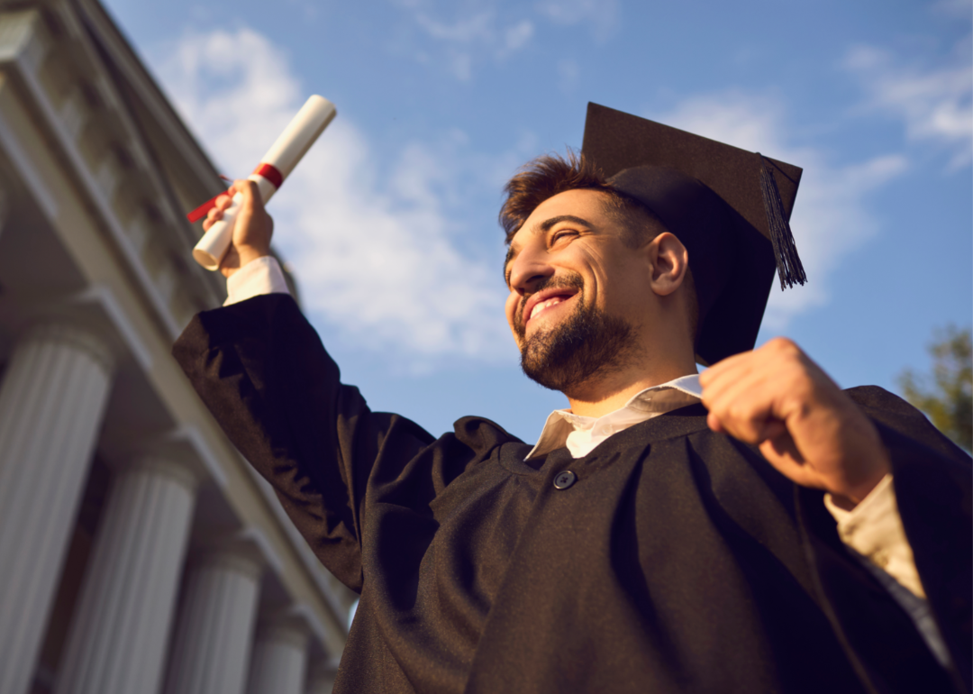 College graduate in black cap and gown holding up diploma and smiling.