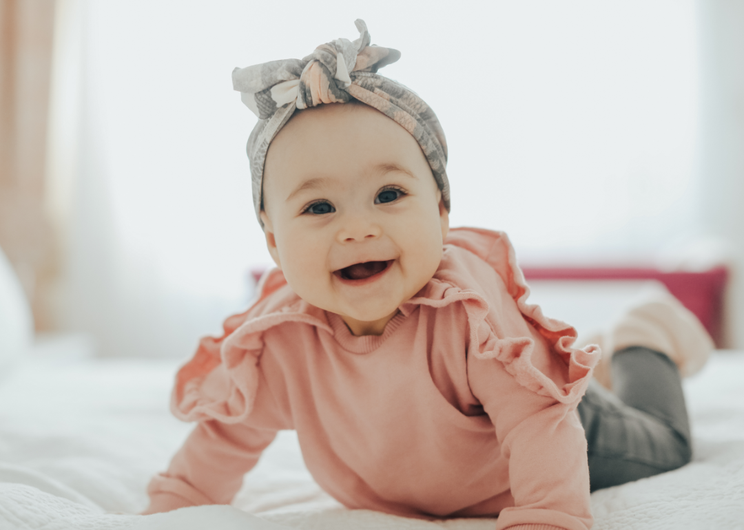 A happy baby in a pink ruffle shirt and headband crawling on the bed and smiling.