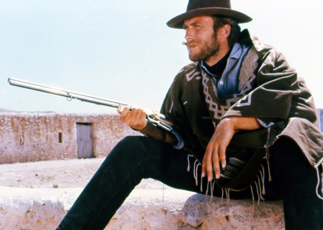 American actor Clint Eastwood smoking a cigar, wearing a brown hat and poncho, taking aim with a rifle in a publicity portrait issued for the film, "A Fistful of Dollars"