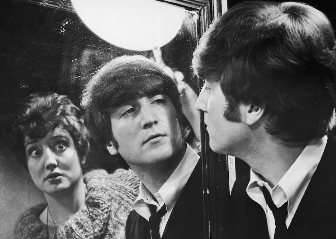 John Lennon from the rock group The Beatles looks at his reflection in a mirror in a still from director Richard Lester's mockumentary 'A Hard Day's Night,' with actor Anna Quayle standing behind him.
