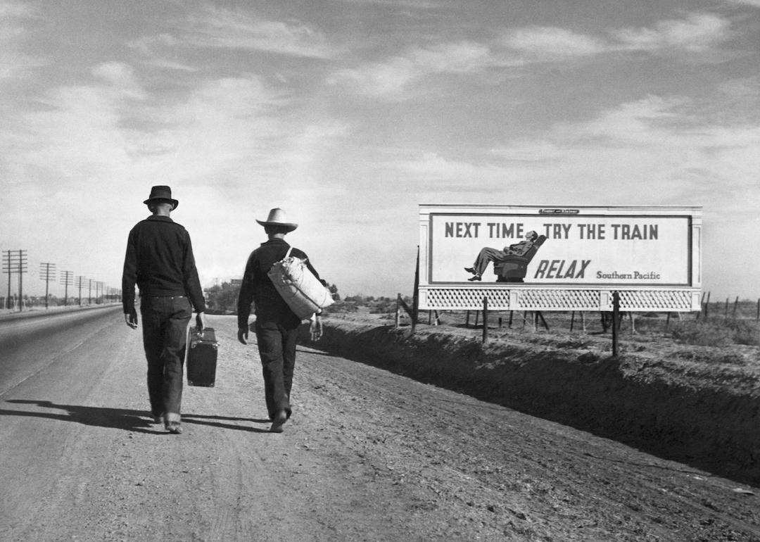 Two Dust Bowl refugees walk along a highway towards Los Angeles. passing by a billboard imploring them 'Next Time Try the Train - Relax.'
