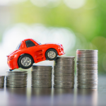 A red toy car rests on stacks of coins that steadily grow taller, signifying a rise in costs.