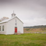 A small white wood-paneled church in a rural field beside a graveyard