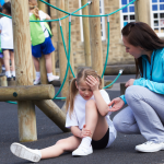 A teacher helps a school-age girl who has fallen hurt herself on the playground.