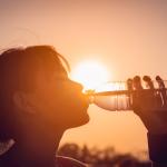 Person with bangs and a ponytail drinks from a water bottle with the sun blazing behind them.
