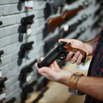 Hands holding and checking out a hand gun in gun shop, a wall of guns in the background