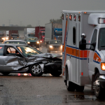 An ambulance pulls up to the scene of a traffic collision where a silver sedan has been totaled