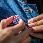 Hands attaching an "I Voted" pin to a denim jacket.