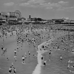 Elevated view along the coastline showing a crowded beach and ocean in Coney Island, New York, circa 1963. Luna Park can be seen in the background. 