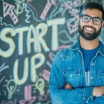A young Indian man with a beard and glasses, smiling, stands in front of a sign reading "START UP"