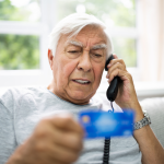 An older man is on the phone, looking distressed, holding up his credit card