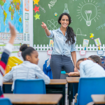 A young female grammar school teacher stands at the front of the class and raises her hand to call on a student