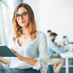 A young female professional wearing glasses holds a tablet in an office setting