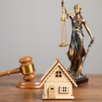 On desk sit a small wooden house, a judge's gavel, and a bronze statue of Lady Justice