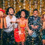 Five party guests in colorful outfits blow glitter confetti into the air.