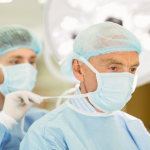 A younger nurse help secure an older surgeon's mask.