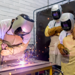 two college-age students watching their classmate practice welding