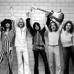 The members of Aerosmith— Steven Tyler, Tom Hamilton, Joe Perry, Brad Whitford, and Joey Kramer—pose backstage with a guitar in 1976.